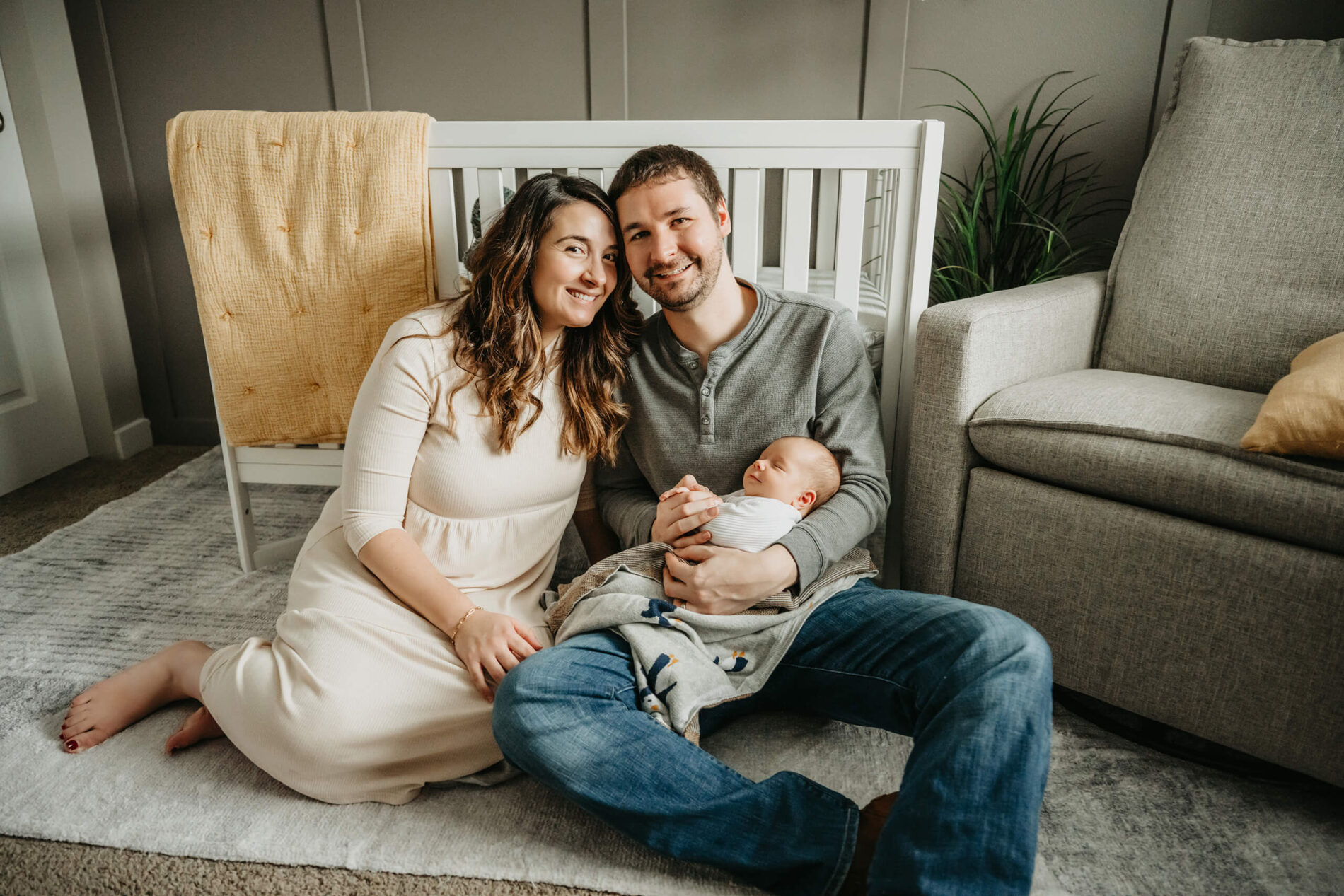 Husband and wife sitting on a floor in a nursery, holding their sleeping newborn son