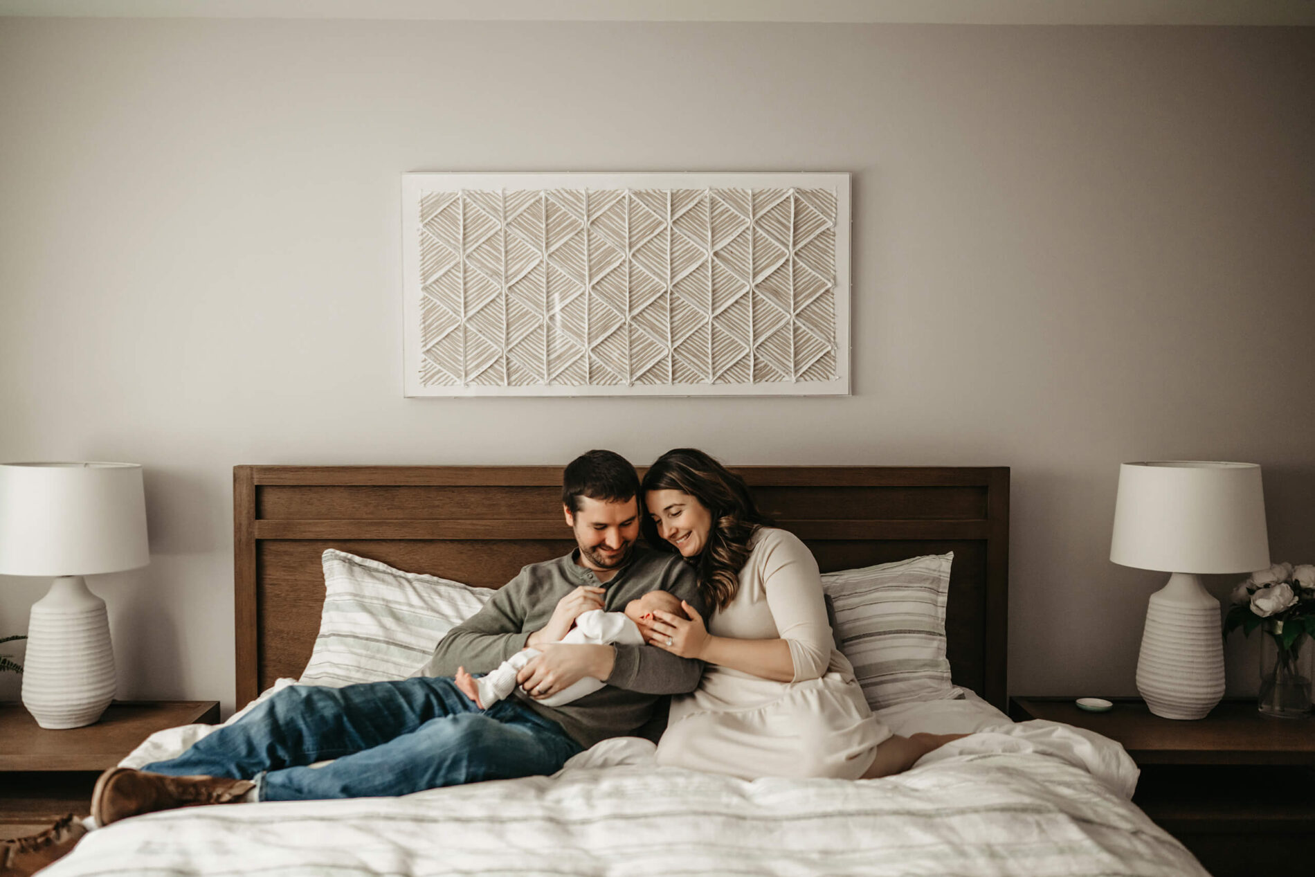 A newborn in-home lifestyle photo of mom and sitting on their bed, holding their newborn son, looking at with joy and happiness