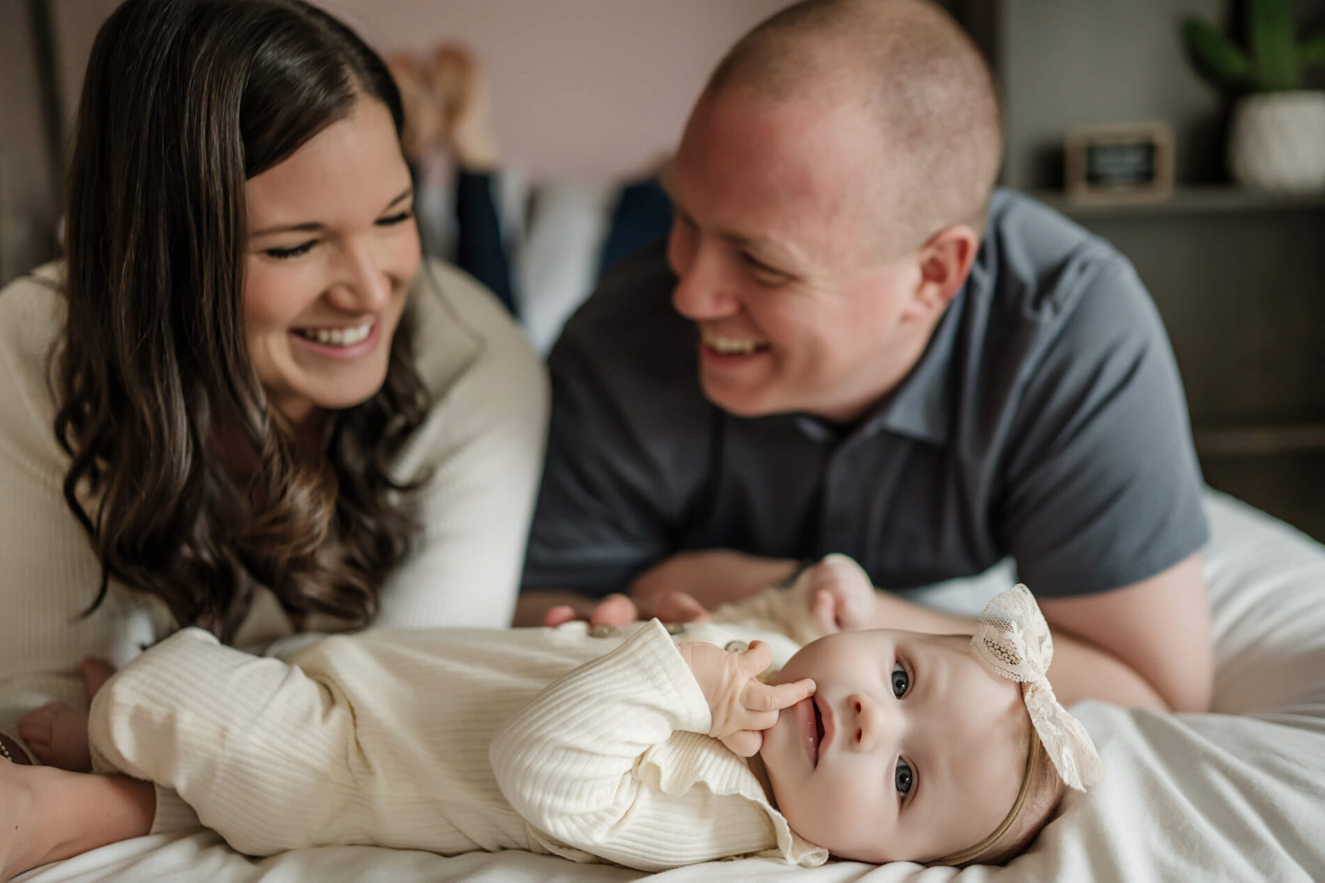 Husband and wife lying on their bed smiling and talking with their baby daughter sucking on her finger lying in front of them