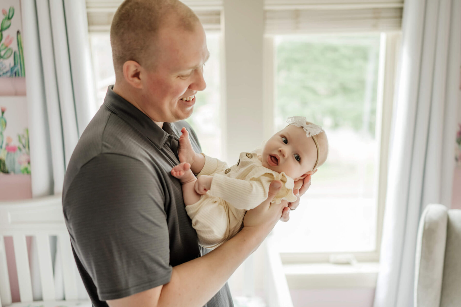 Dad lovingly holding his newborn daughter in his arms in front of a large window in a nursery
