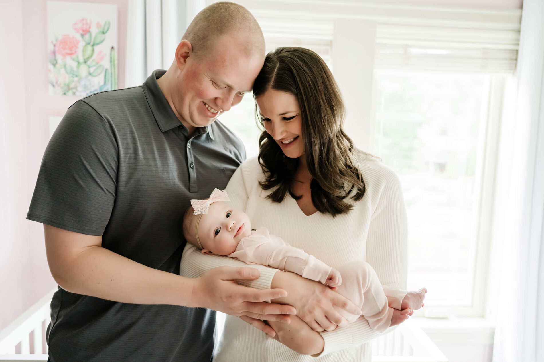 Woman hugged by her husband and holding her newborn girl standing in front of a window