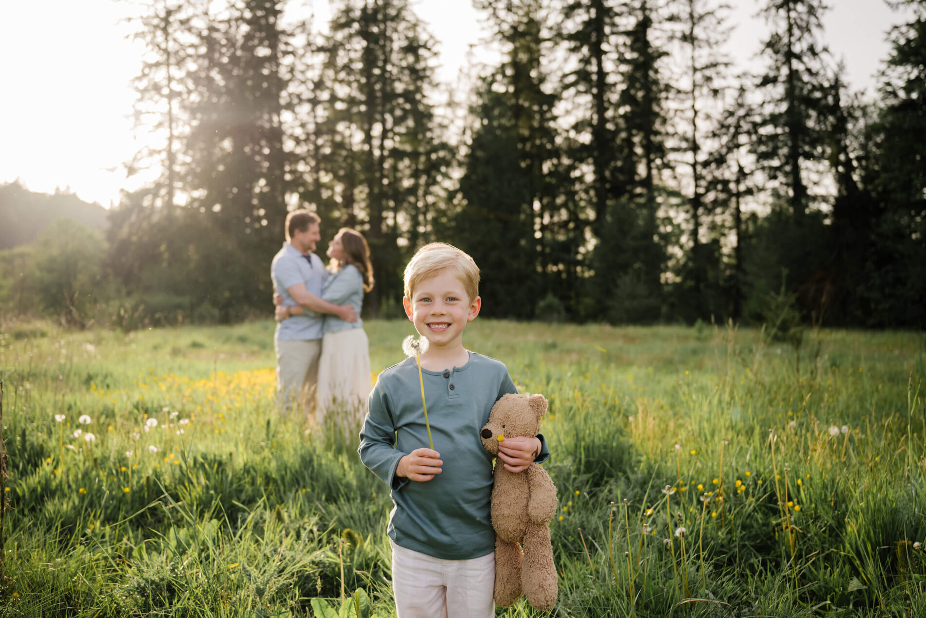Young child holding a teddy bear and dandelion in a sunlit field, with parents softly blurred in the background.