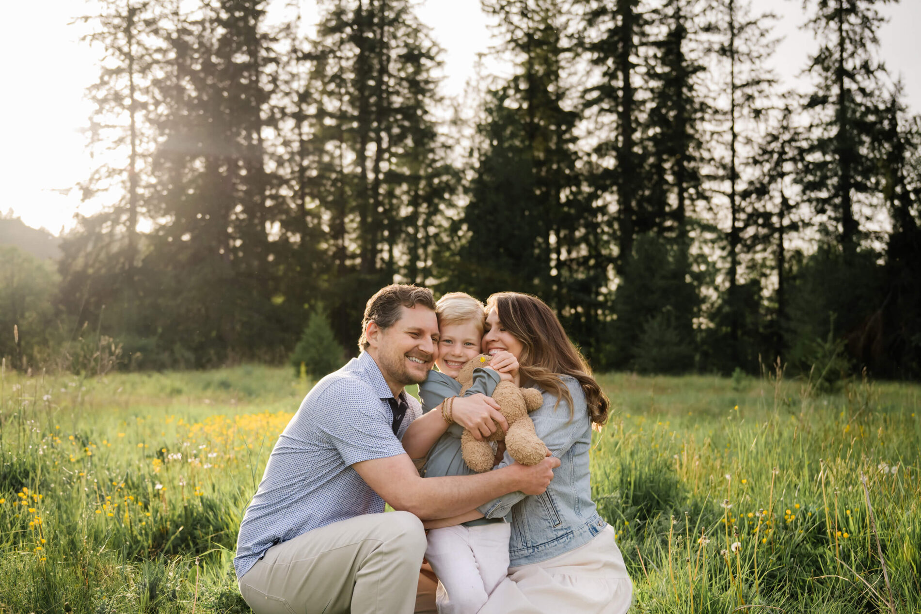 Family of three cuddling together in a grassy meadow with yellow wildflowers and evergreen trees behind them.