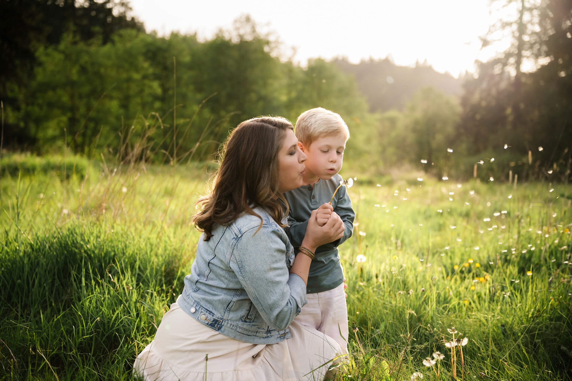 Mother and child blowing dandelion seeds together in a grassy field during golden hour.