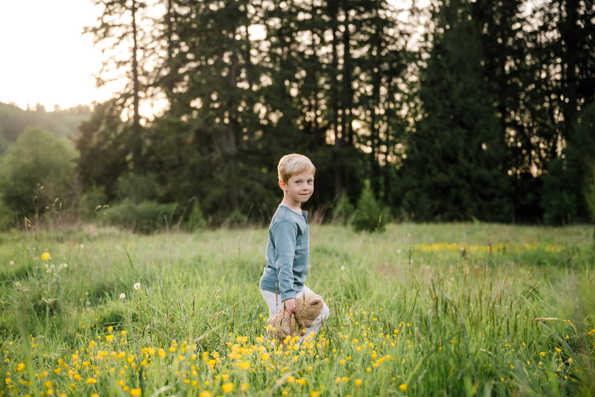 Young child walking through yellow wildflowers while holding a teddy bear in a sunlit meadow.