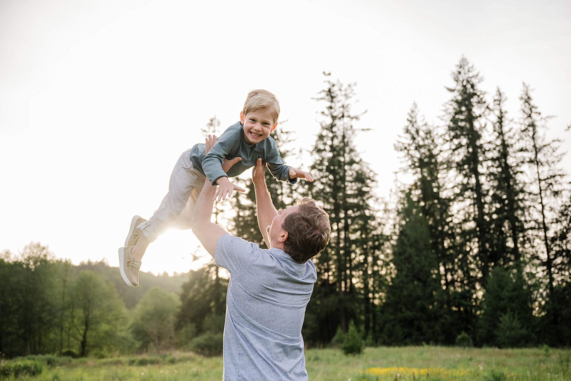 Father lifting his child into the air in a sunny meadow with evergreen trees in the background.