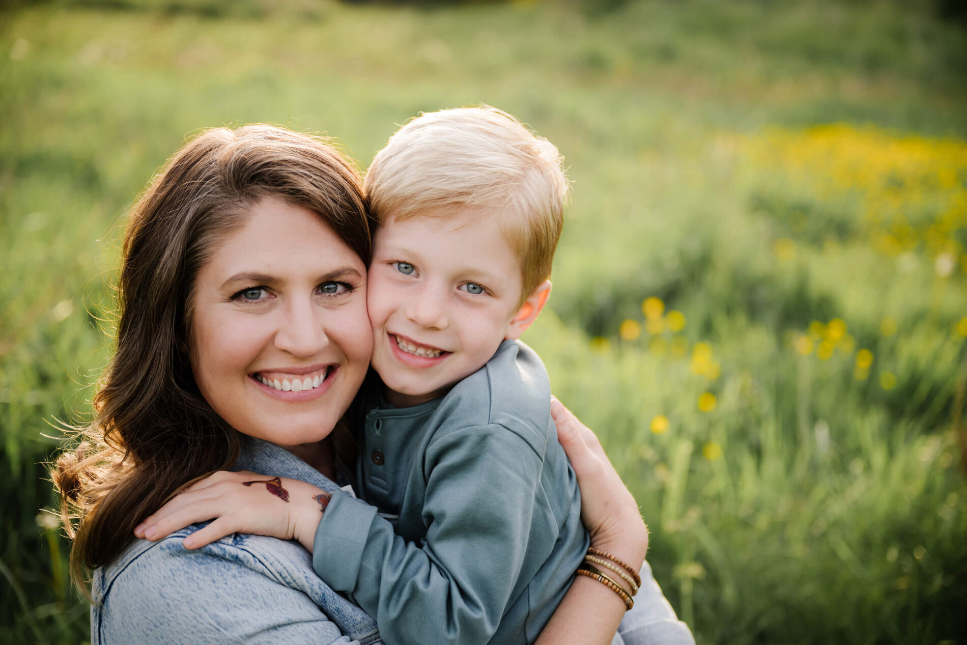 Mother holding her child close for a smiling portrait in a green field with soft yellow wildflowers behind them.
