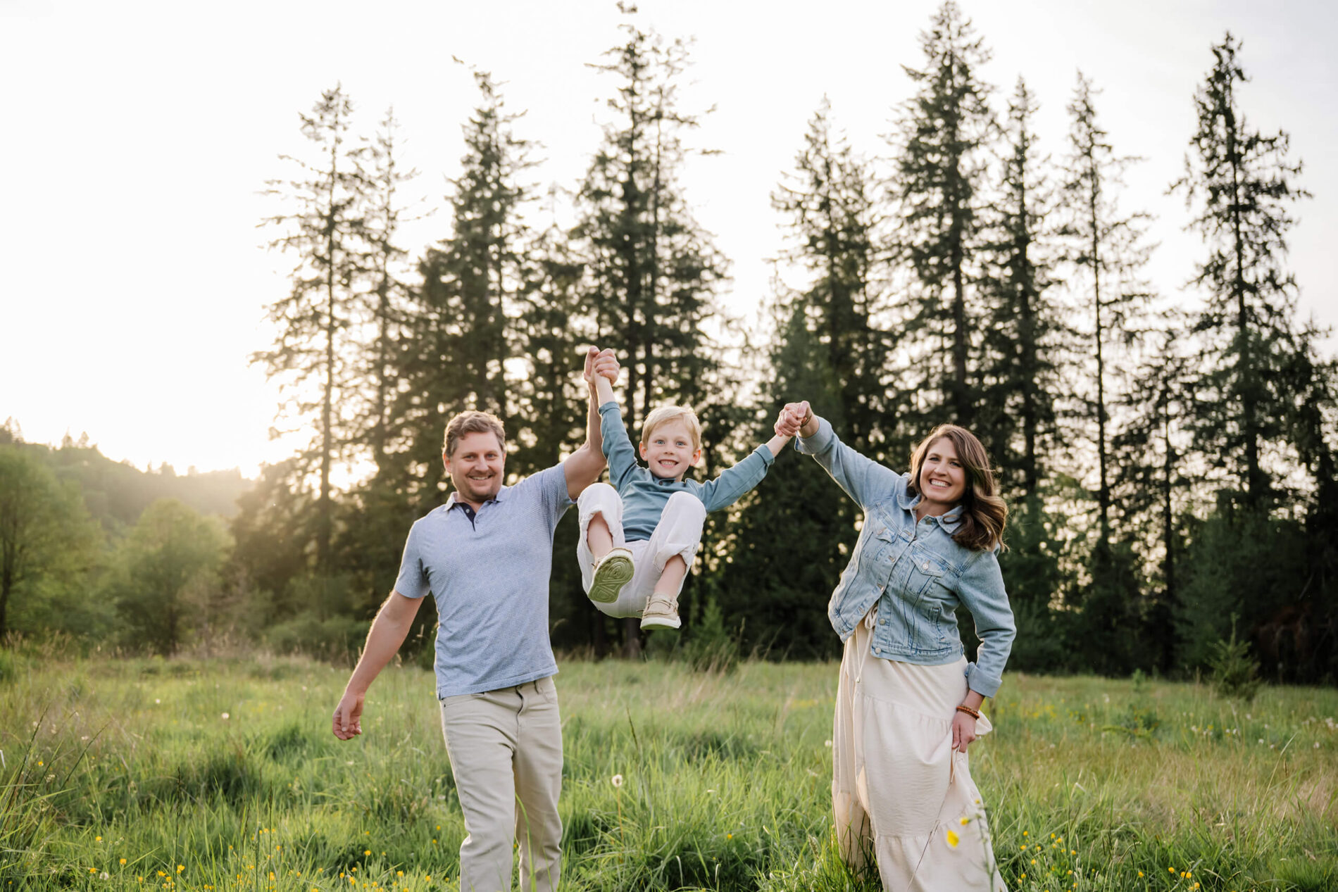 Parents holding their child’s hands and lifting him between them during an outdoor family session in a grassy field.