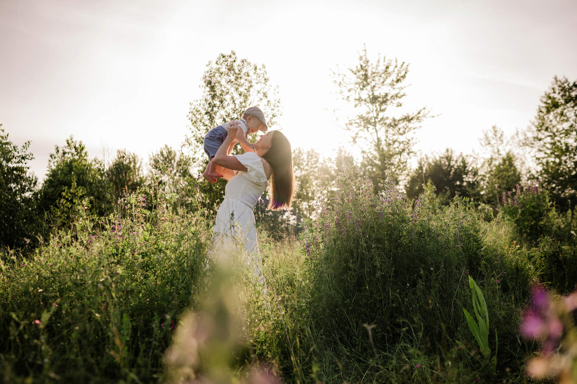 Mother lifting her baby in a wildflower field with soft evening light behind them.