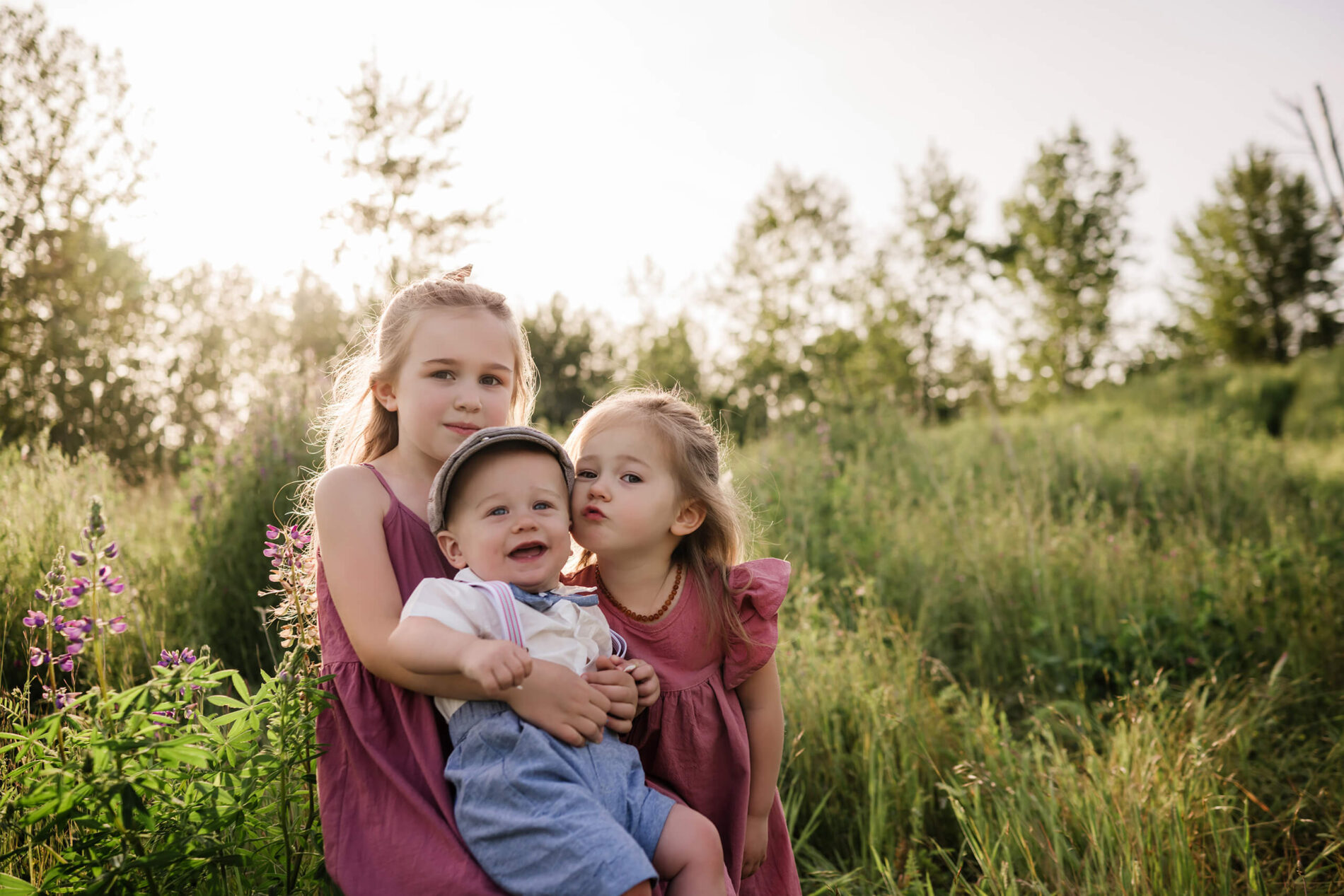 Three young siblings sitting together in a grassy field with purple wildflowers nearby.