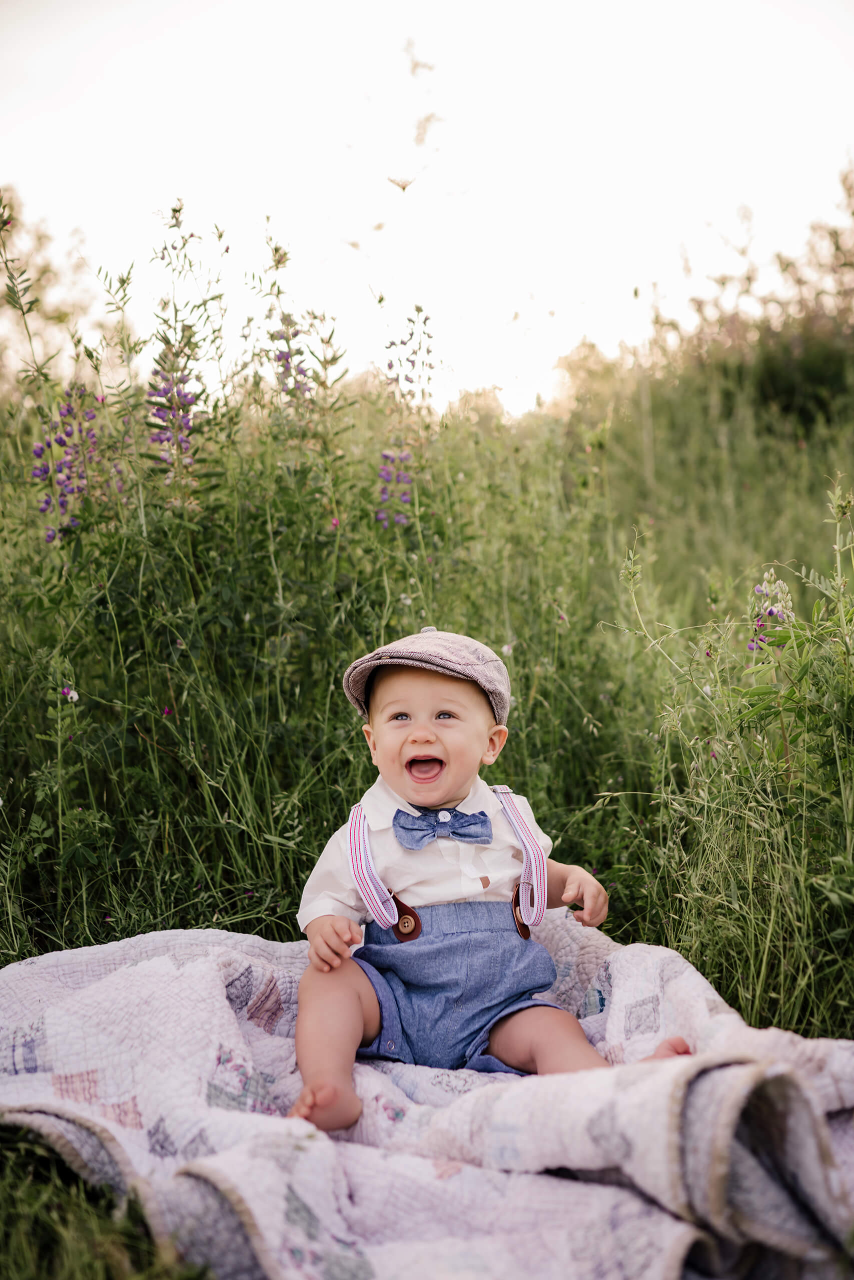 Smiling baby sitting on a quilt in tall grass and purple wildflowers.