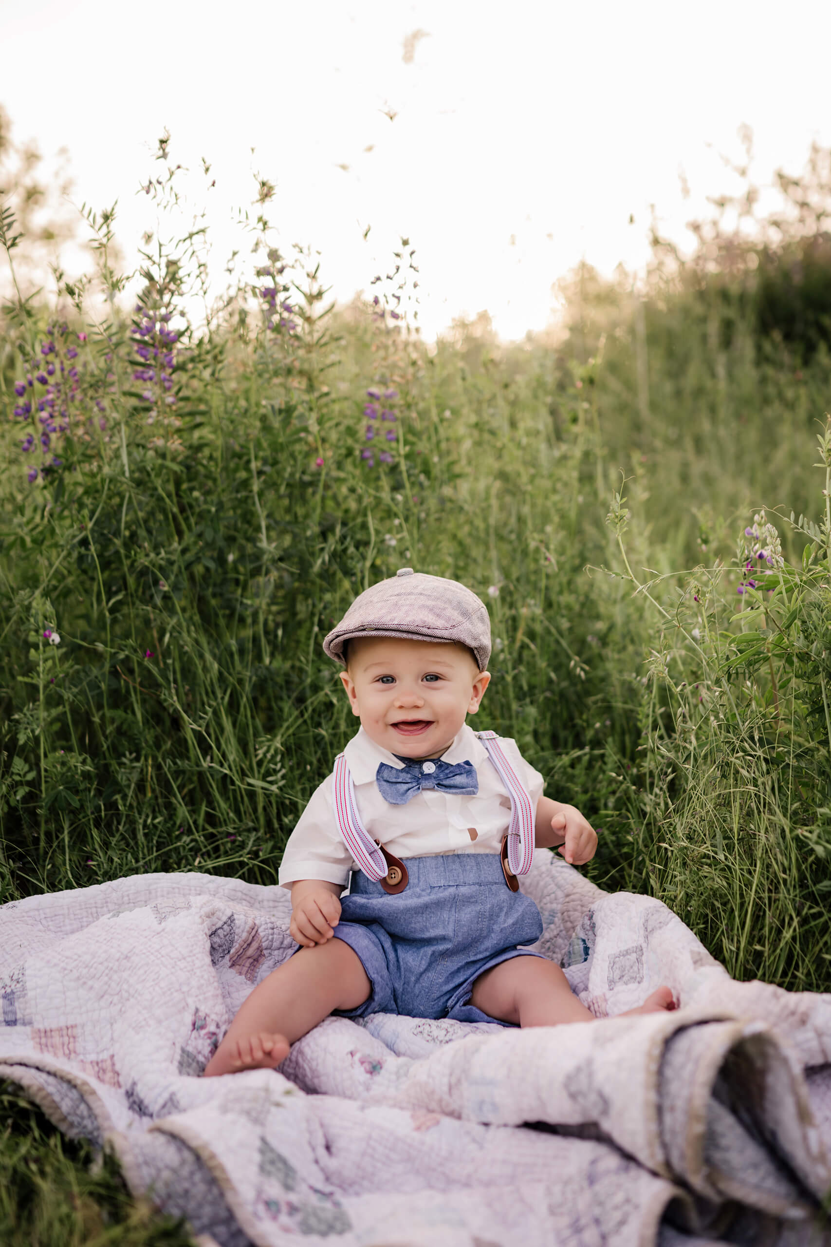 Smiling baby sitting on a quilt in tall grass with purple wildflowers behind him.