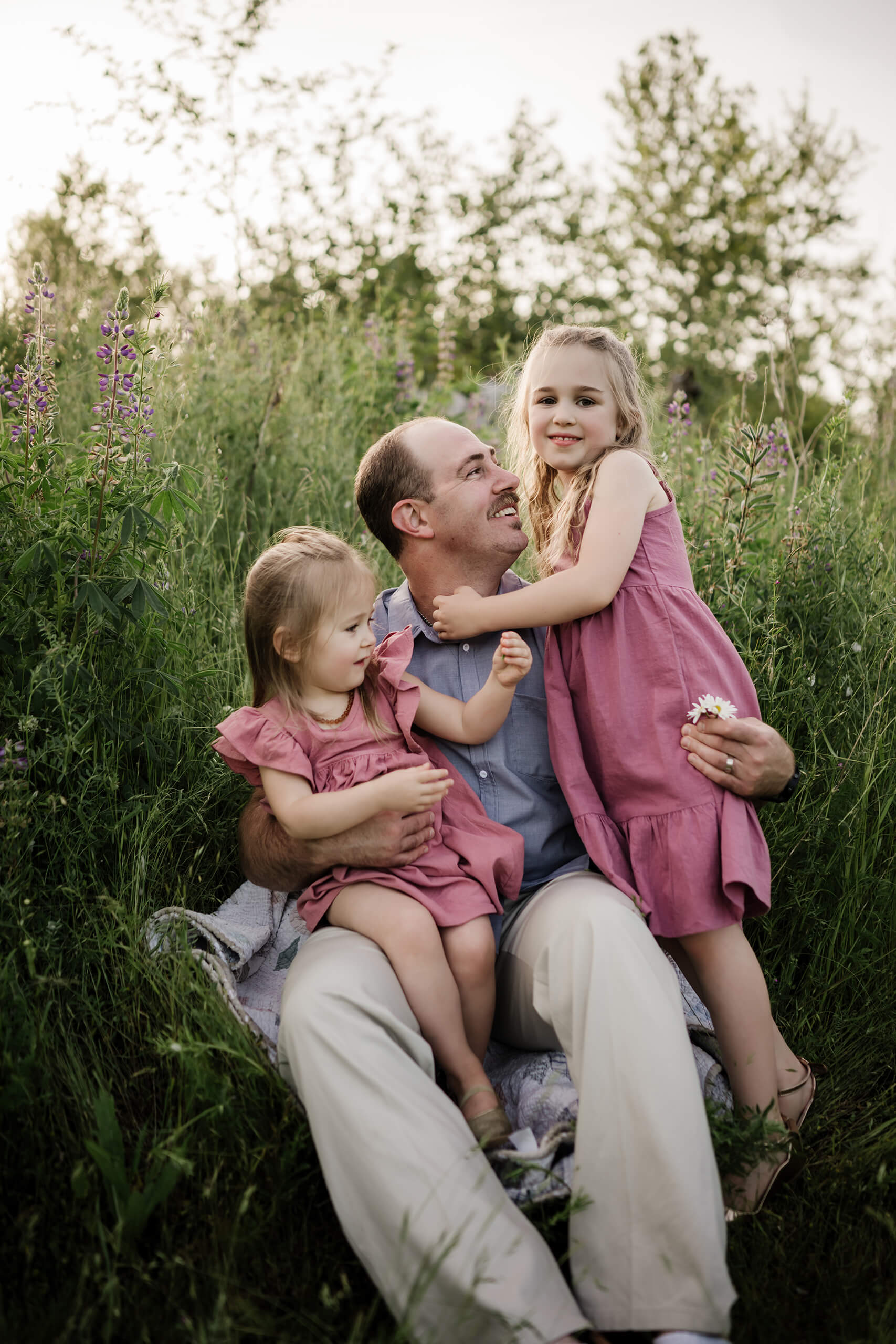 Father sitting with his two daughters in a grassy wildflower field.