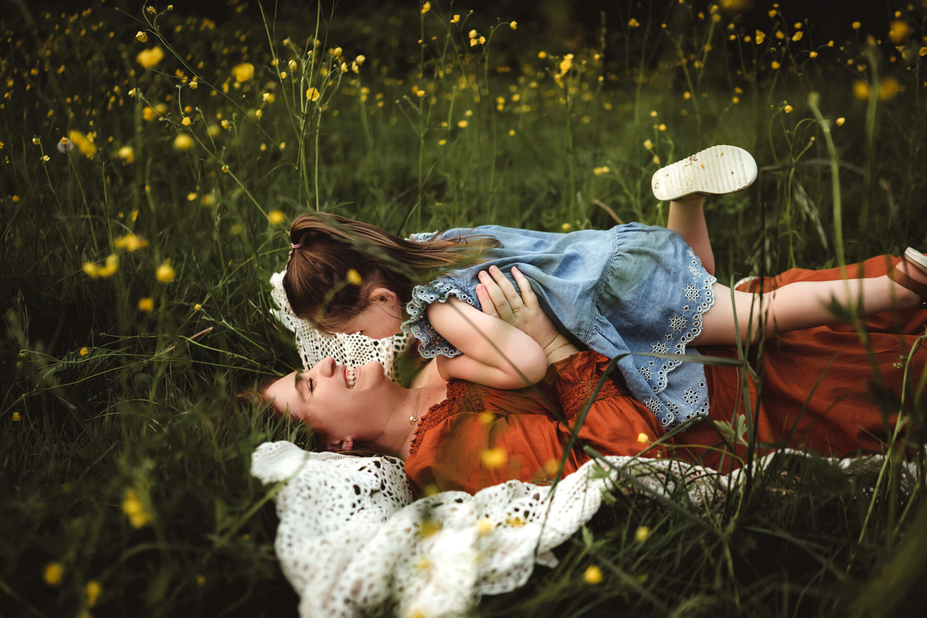 Mom lying on the grass in a wild flower meadow with her young girl on top of her