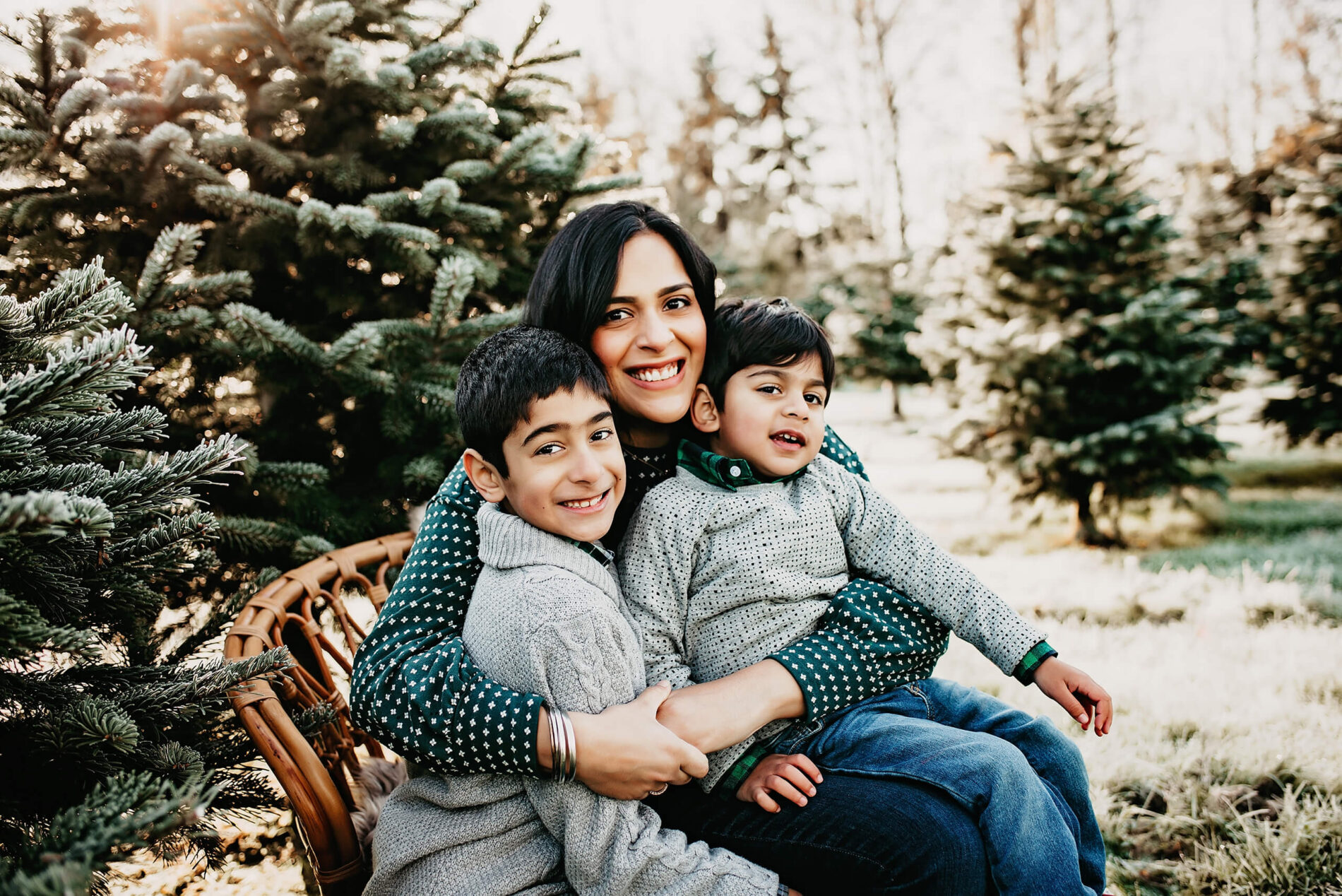 Mom sitting on a wooden chair, holding her two sons on her lap, with Christmas trees in the background