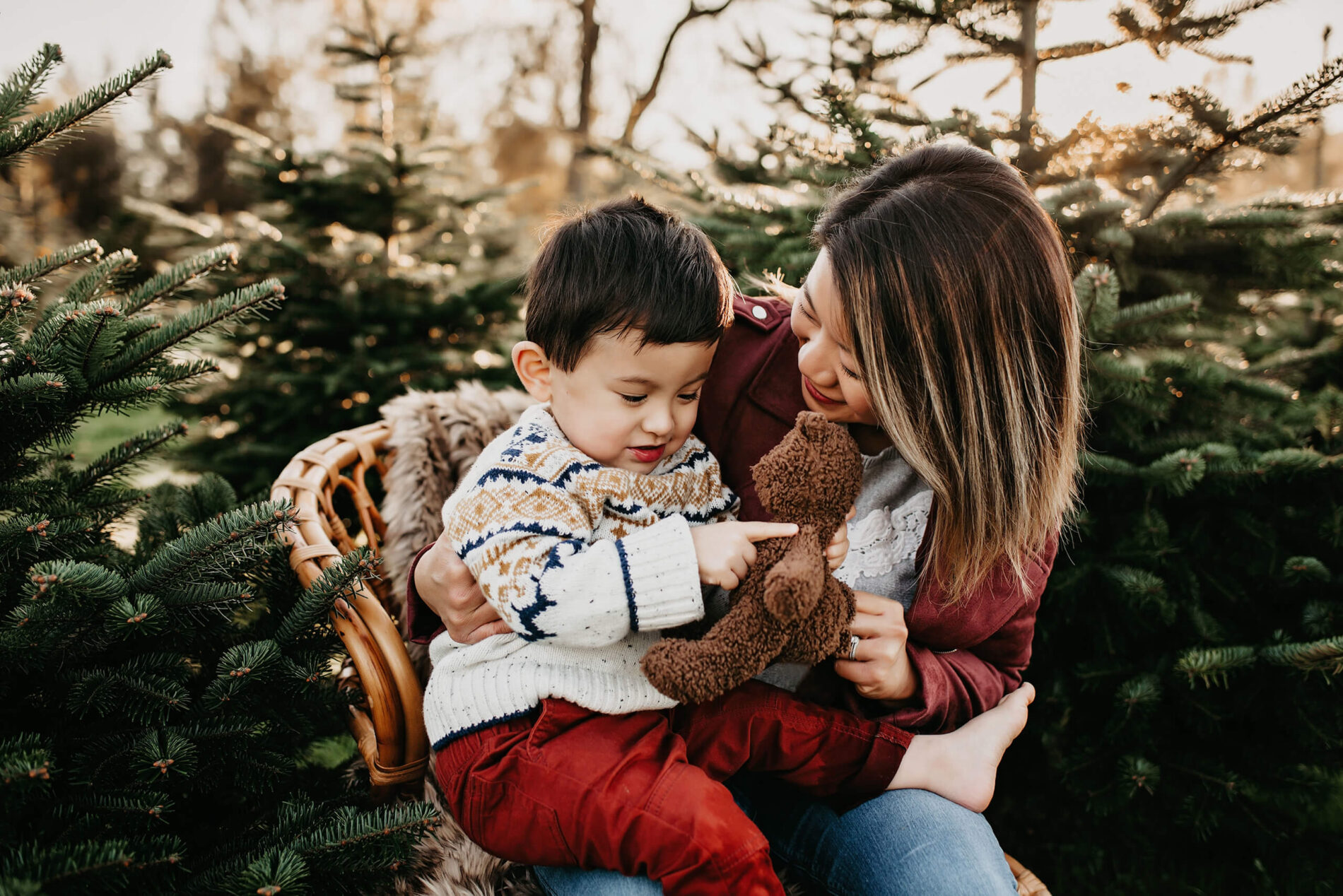 Mom sitting on a wooden chair at a Christmas tree farm, with her son on her lap who is playing with a teddy bear