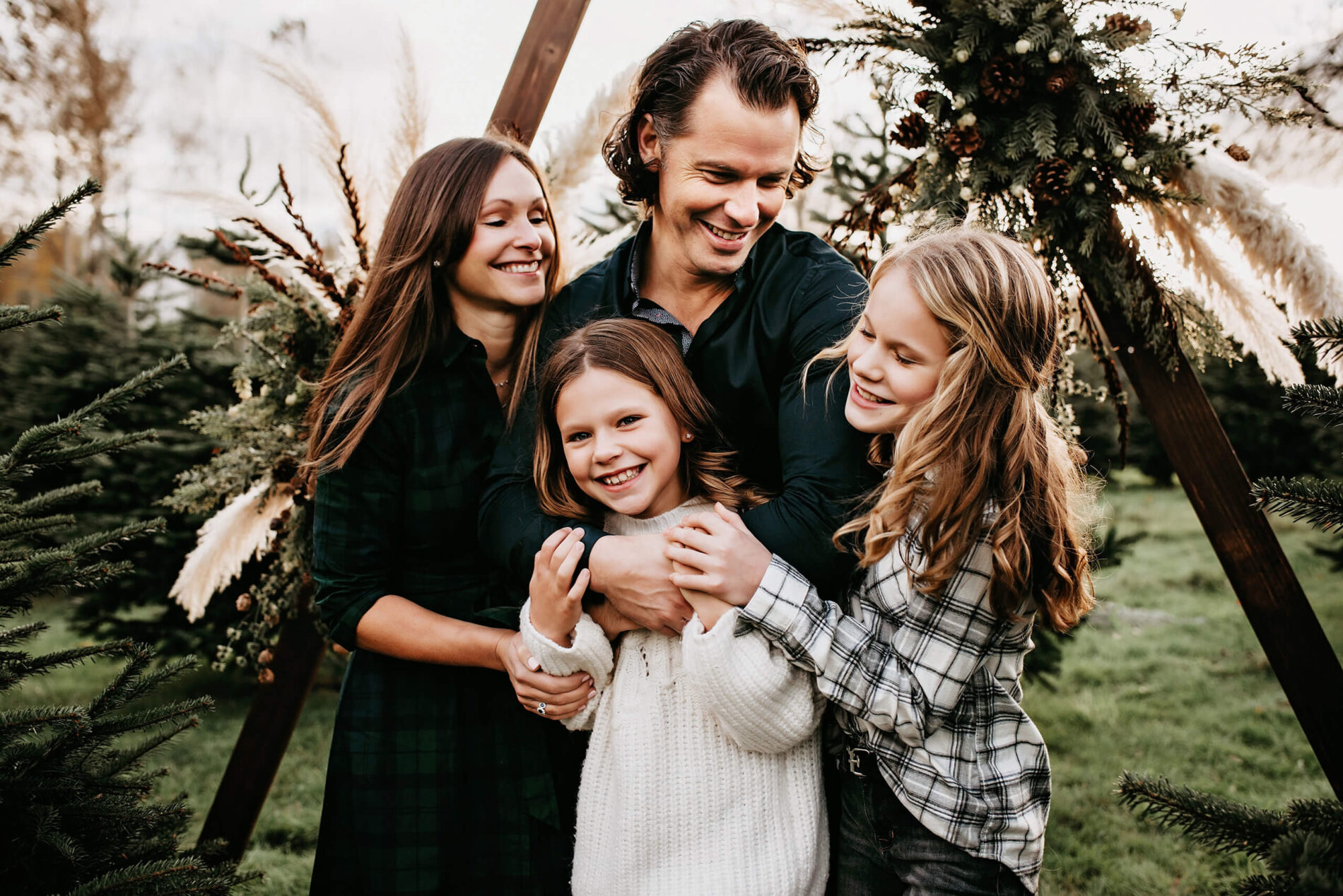 A happy family of mom, dad, and two daughters hugging at a Christmas tree farm