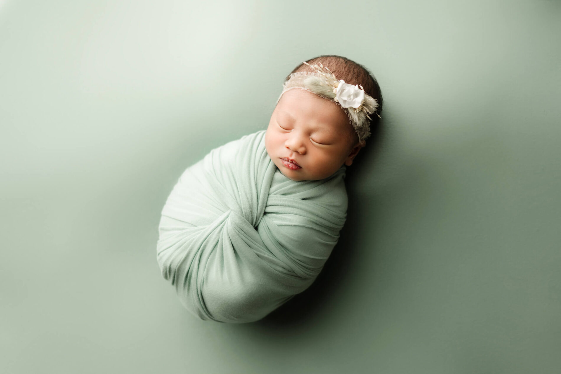 Peacefully sleeping newborn girl during a photo shoot wearing a floral headband and wrapped in a green blanket