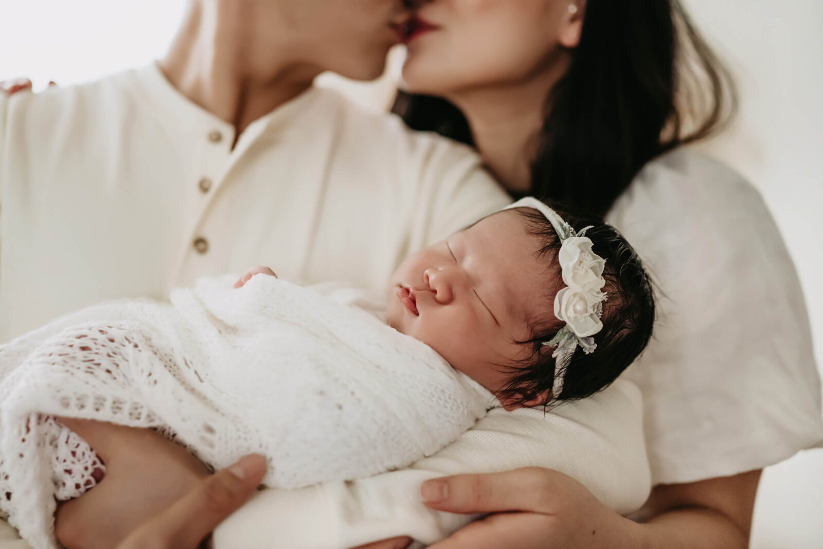 Close-up of a newborn girl, wrapped in white blankets and sleeping, while parents are kissing in the background