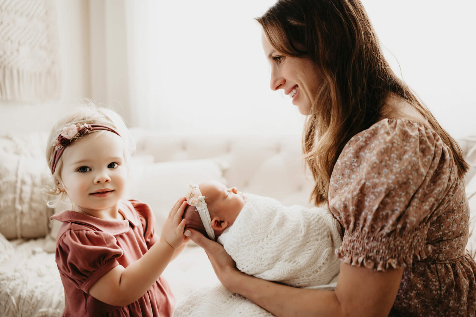 Mom holding her newborn girl and smiling, while her other daughters is touching the newborn