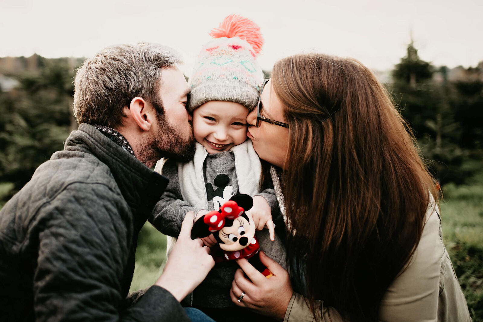 Mom and dad kissing their daughter from both sides at a Christmas tree farm