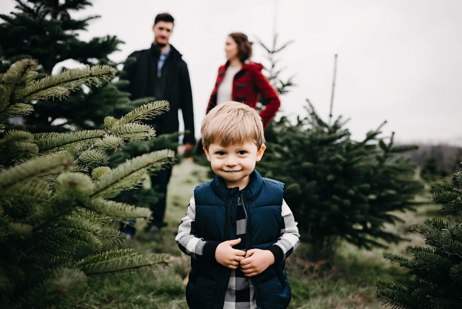 A cute toddler at a Christmas tree farm with parents holding hands blurred in the background