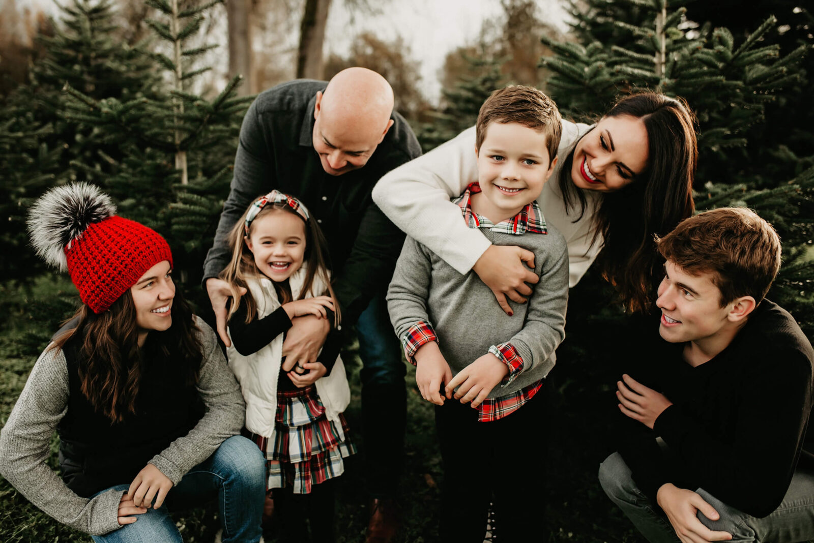 Seattle Christmas tree farm family holiday session with parents and four children laughing among evergreen rows; little girl in plaid dress, brother in gray sweater, teens in winter outfits.