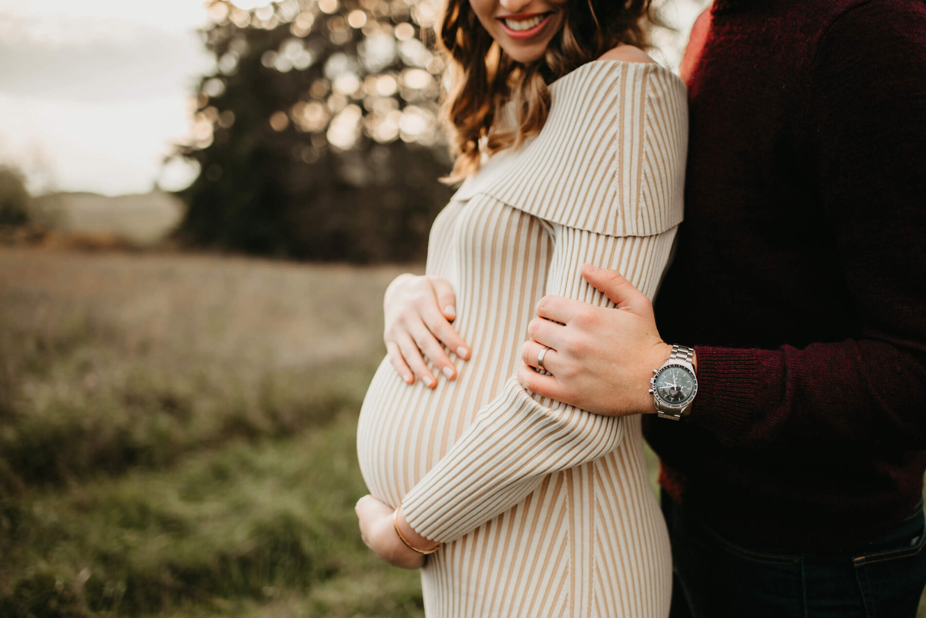 A smiling pregnant woman, lovingly hugged by her husband in a natural setting
