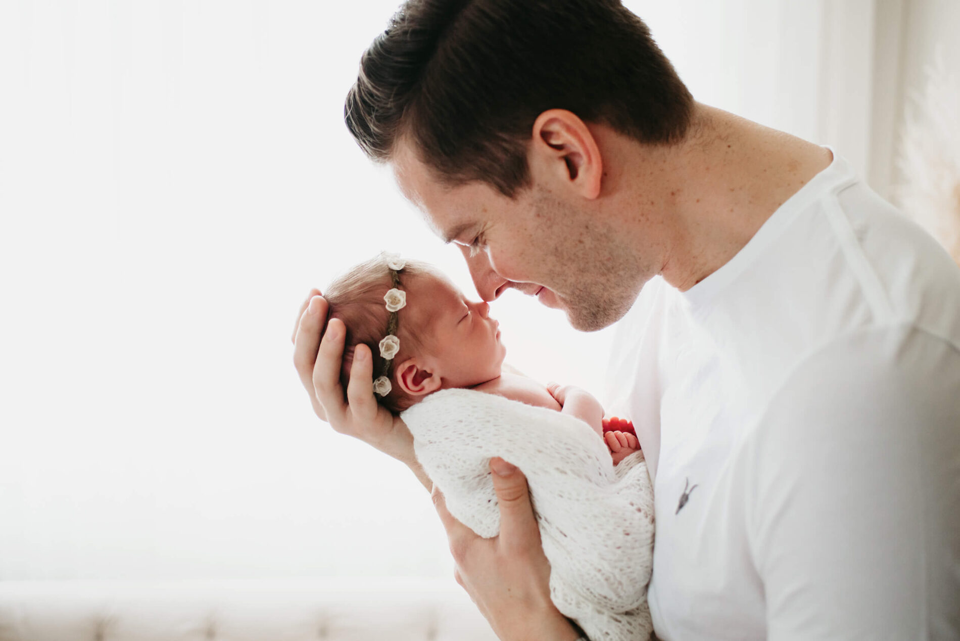 A man, smiling lovingly, holding his newborn daughter close to his face, their noses touching