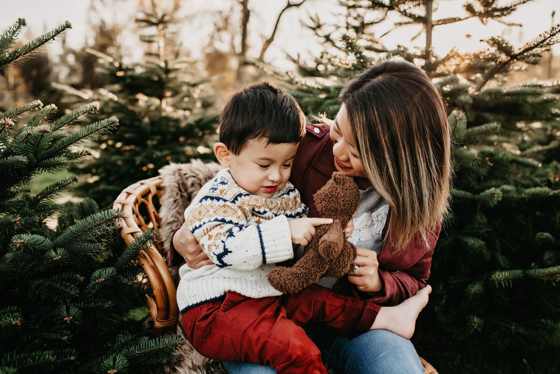 A photo at a Christmas tree farm of a Mom sitting in a wooden chair with her son playing with a teddy bear