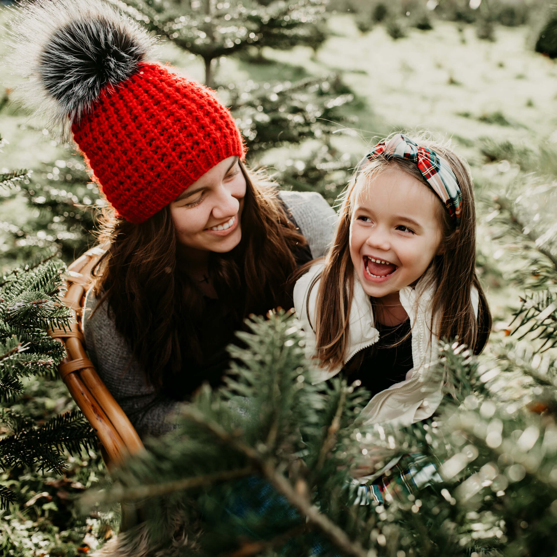An older sister wearing a festive hat with a pom pom posing with her younger sister at a Christmas tree farm