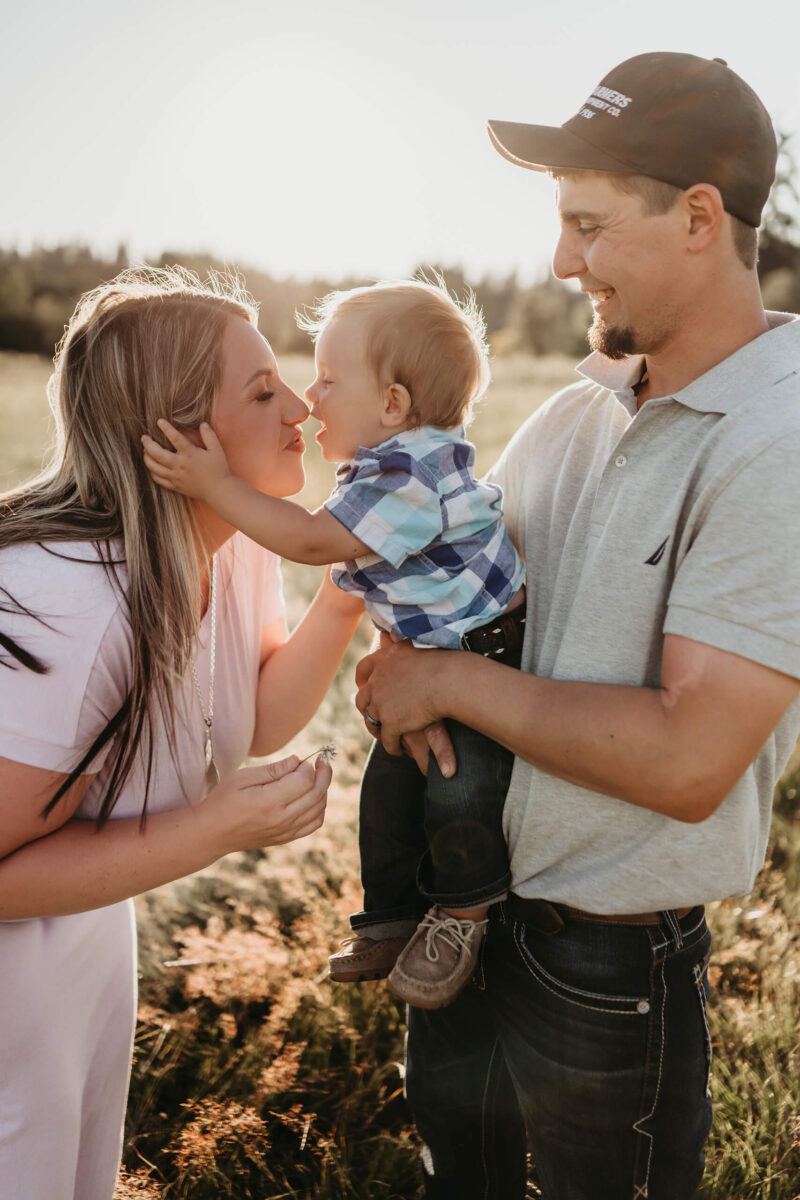 Family photo shoot of a dad holding his toddler son who is reaching over to kiss mom