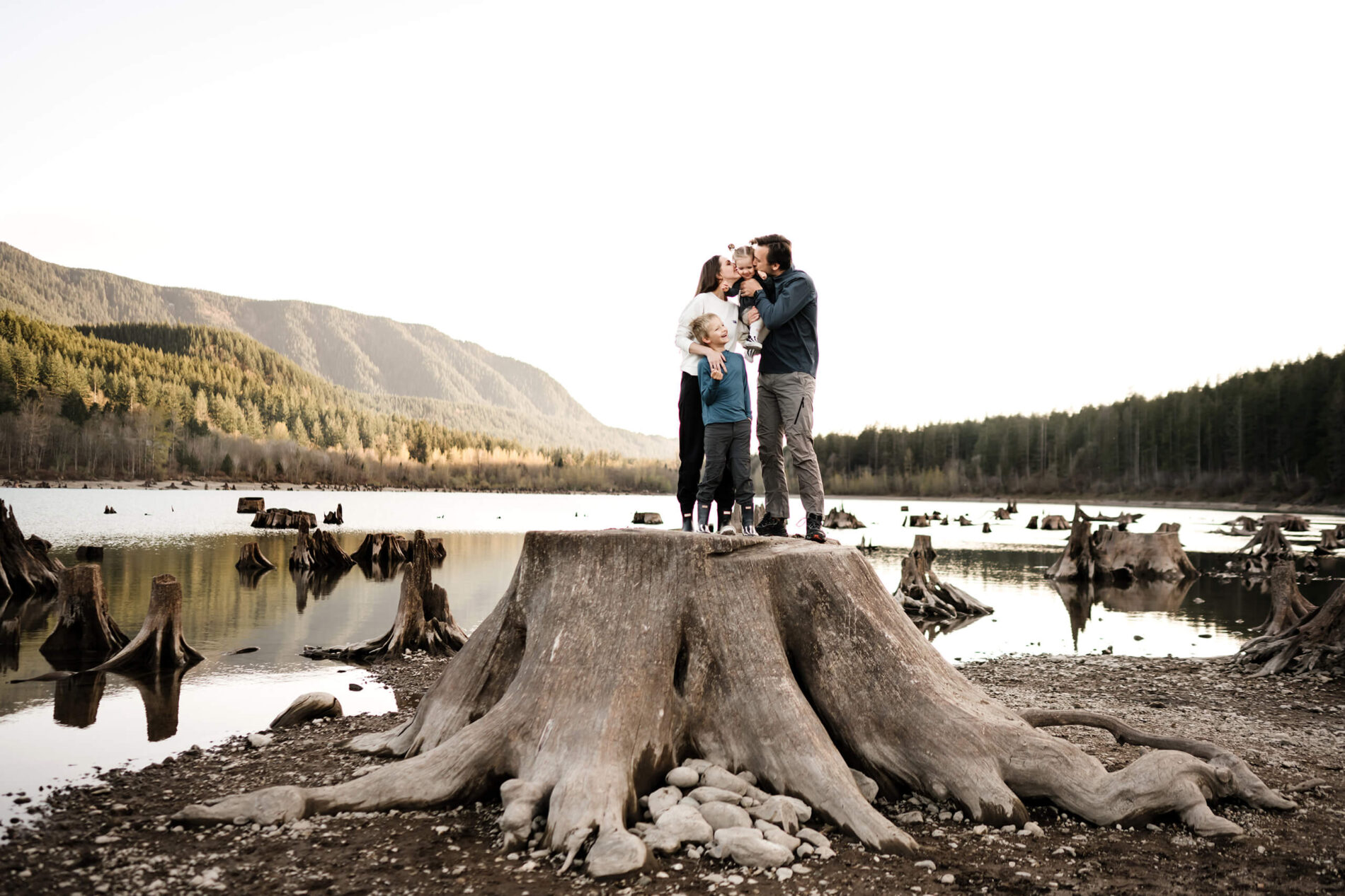 Family of four standing on top of a large tree trunk with stunning lake and mountain views in the background