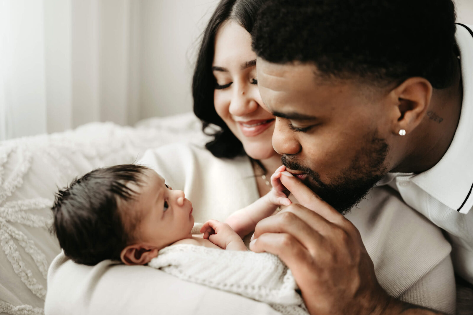 Seattle-area newborn studio session: dad kisses baby’s tiny fingers while mom smiles over their swaddled newborn in a cream knit blanket—soft window light.