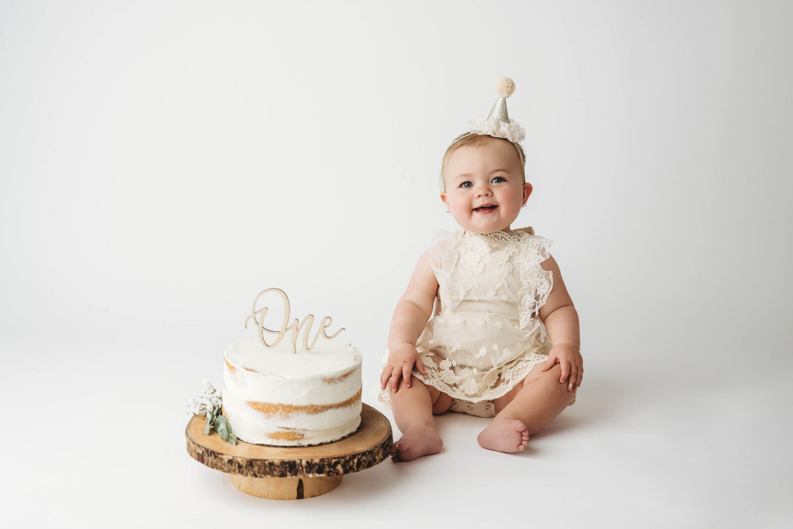 Cake smash photo session of a smiling one year old girl sitting in front of a white cake on a wooden cake stand