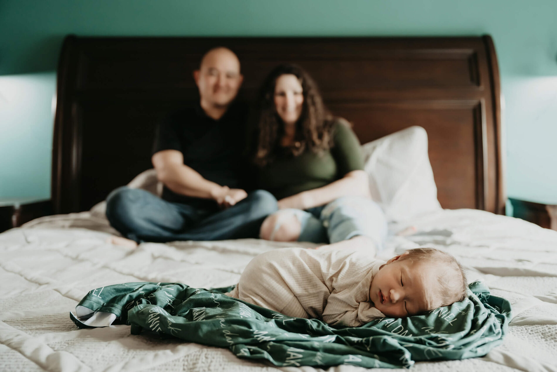 Unique newborn photography with parents: newborn boy lying on parents' bed with mom and dad smiling and blurred in the background