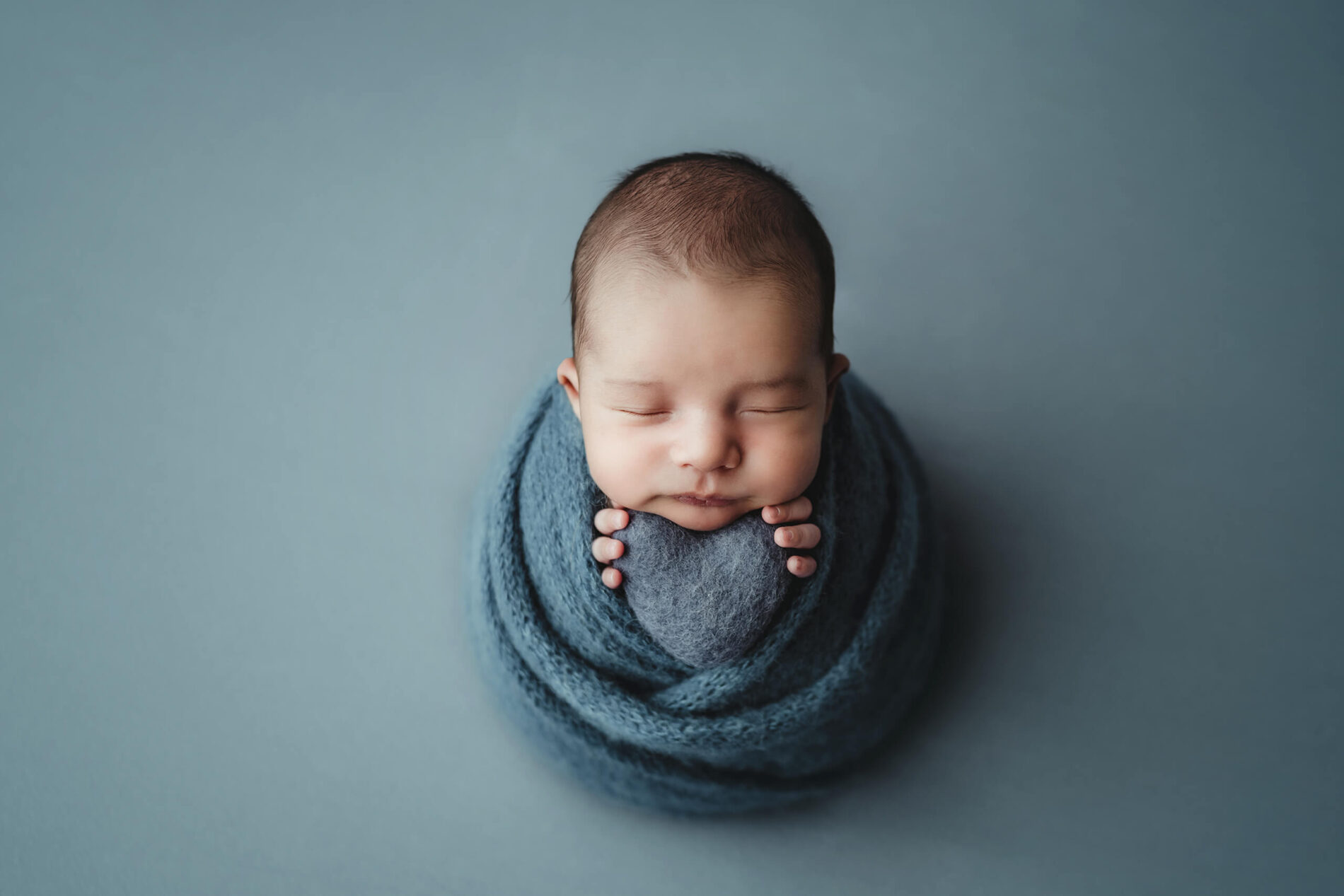 Newborn photography session: cozily-wrapped sleeping newborn boy holding a stuffed heart prop