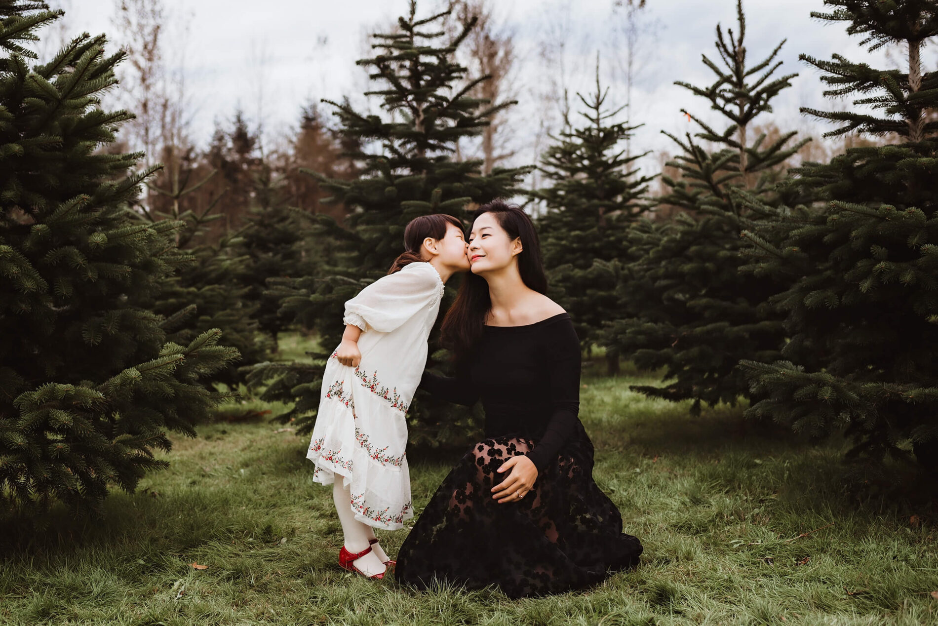 A young girl elegantly dressed kissing her mom at a Christmas tree farm