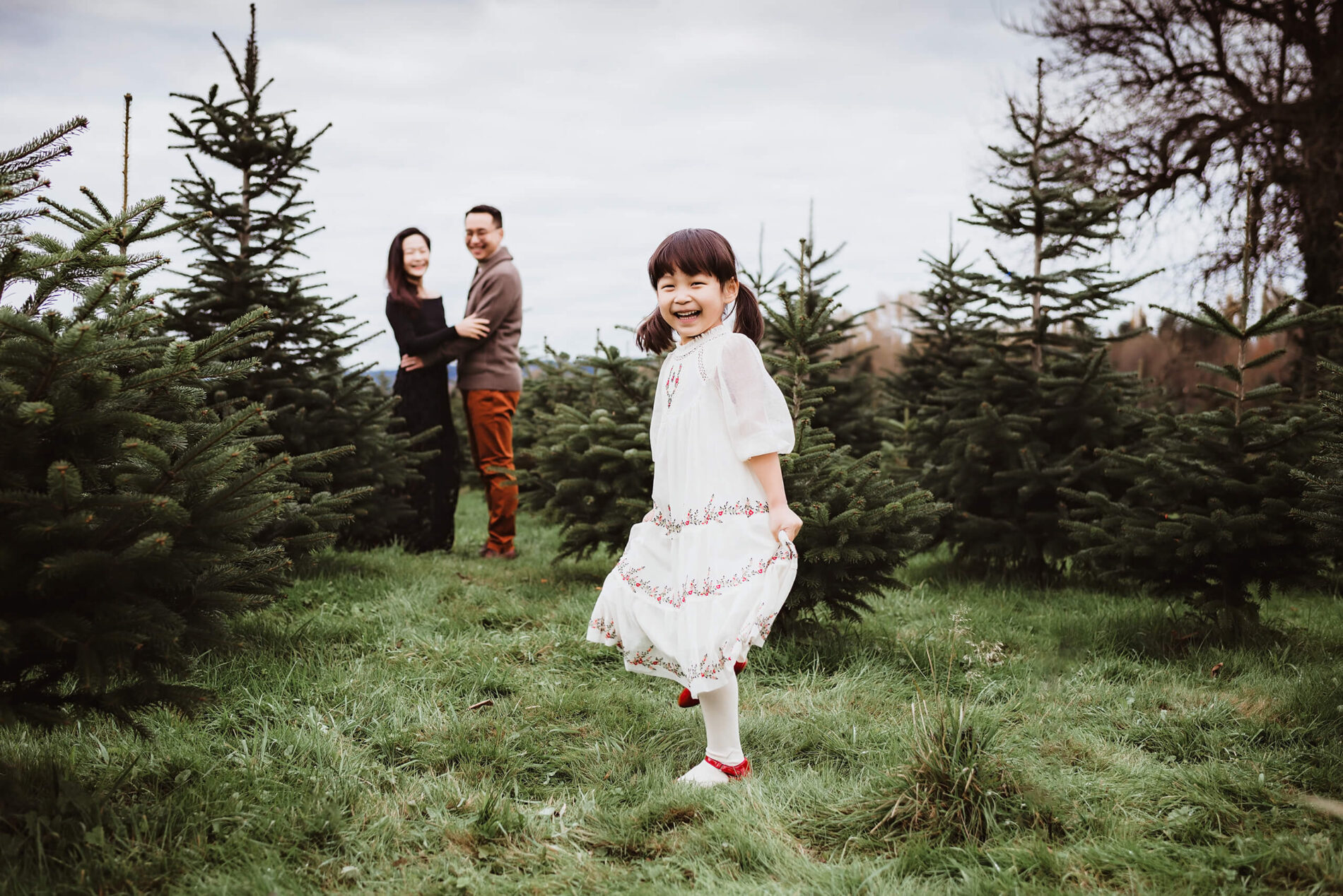 A girl in a beautiful dress twirling at a Christmas tree farm with parents in the background during a photoshoot