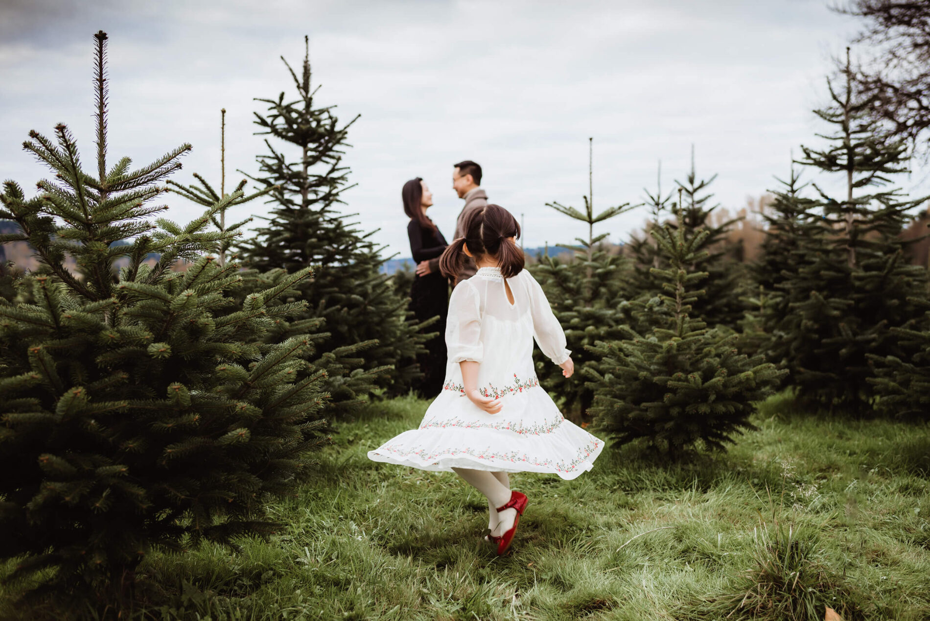 A girl in a beautiful dress twirling at a Christmas tree farm with parents in the background