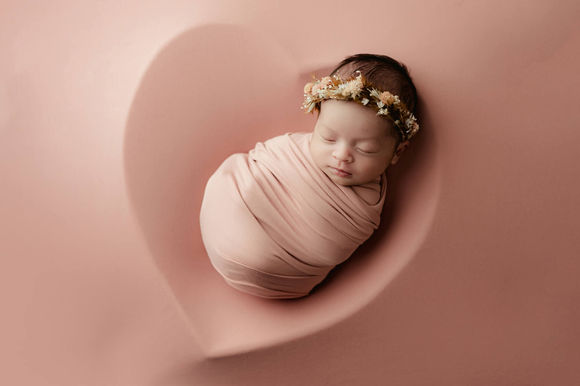 Newborn portrait of a sleeping girl in pink wraps with a floral headband lying in a heart-shaped basket