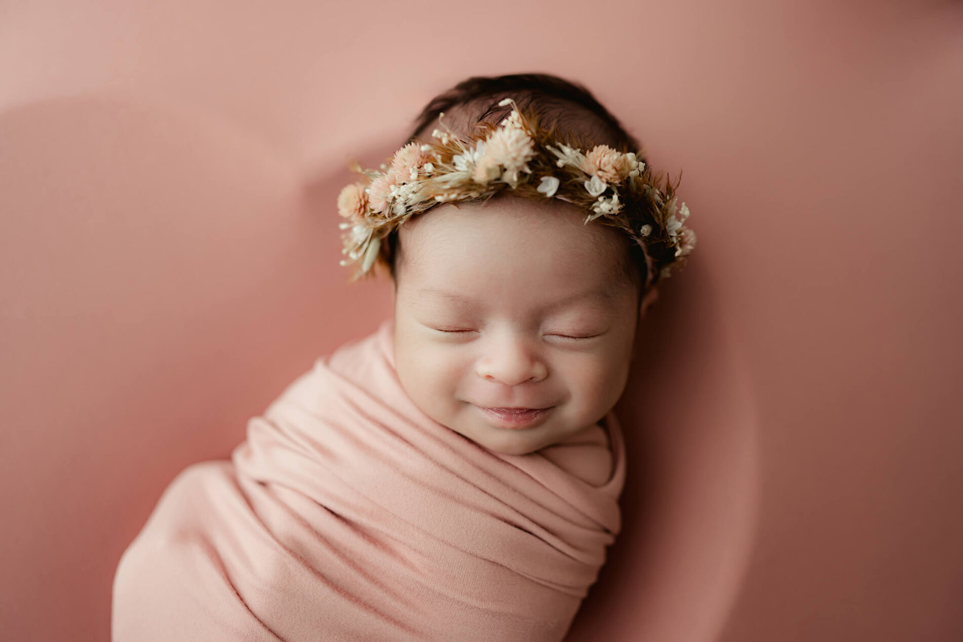Newborn portrait of a sleeping and smiling girl in pink wraps with a floral headband