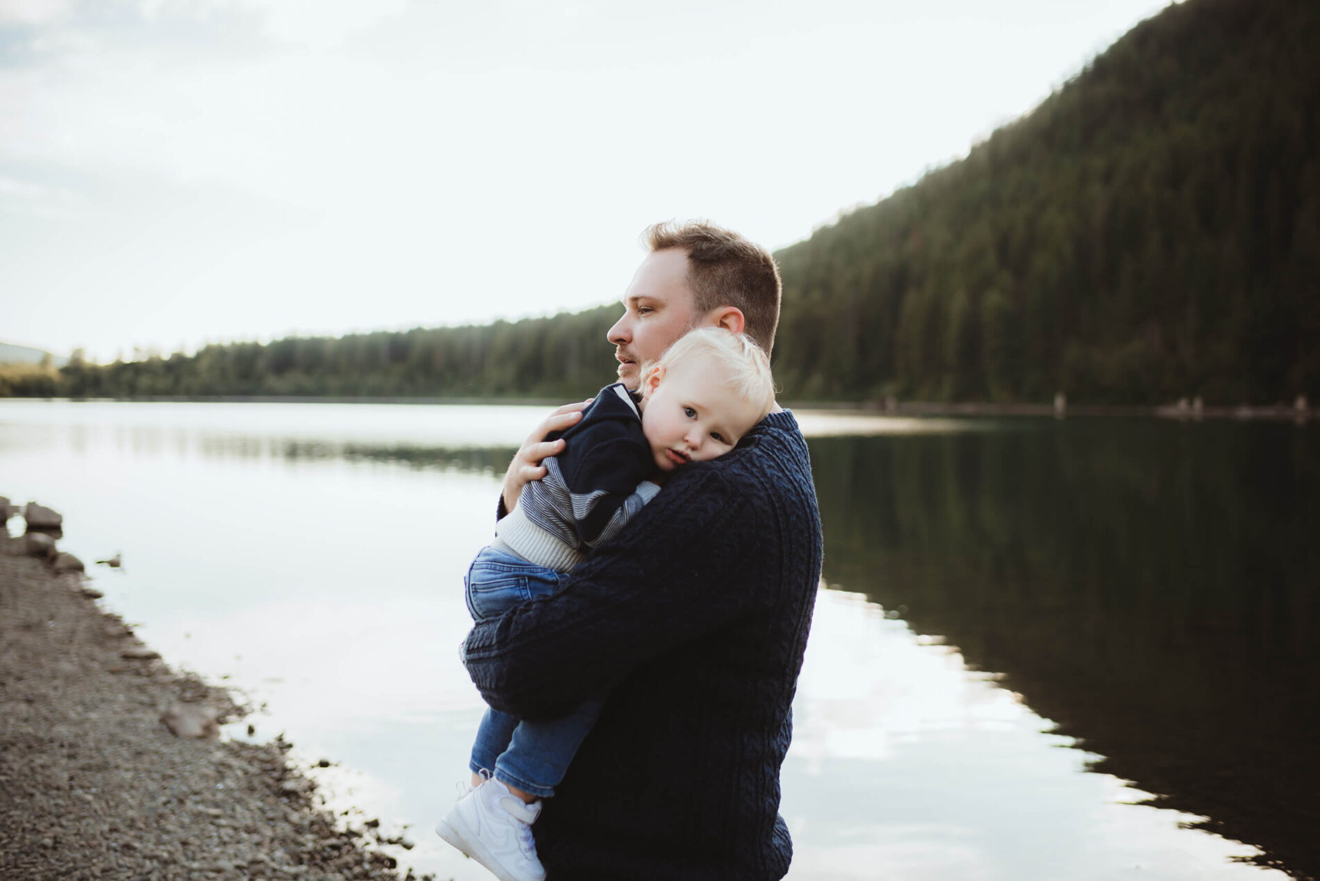 A moment during a family photo shoot of dad lovingly holding his toddler son with lake and mountains in the background