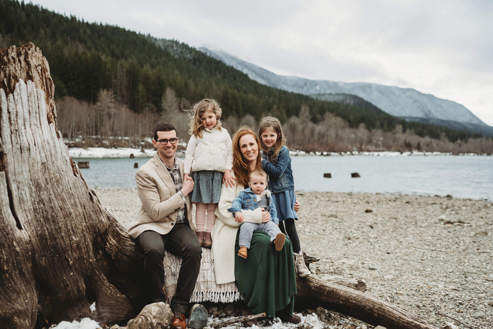 Natural family portrait of mom, dad, with three children, sitting on a large tree stump with lake and mountains in the background