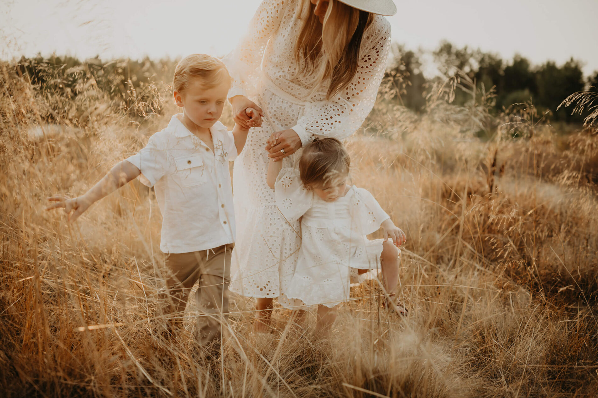 Family photo shoot with mom in white dress walking with son and daughter in a field
