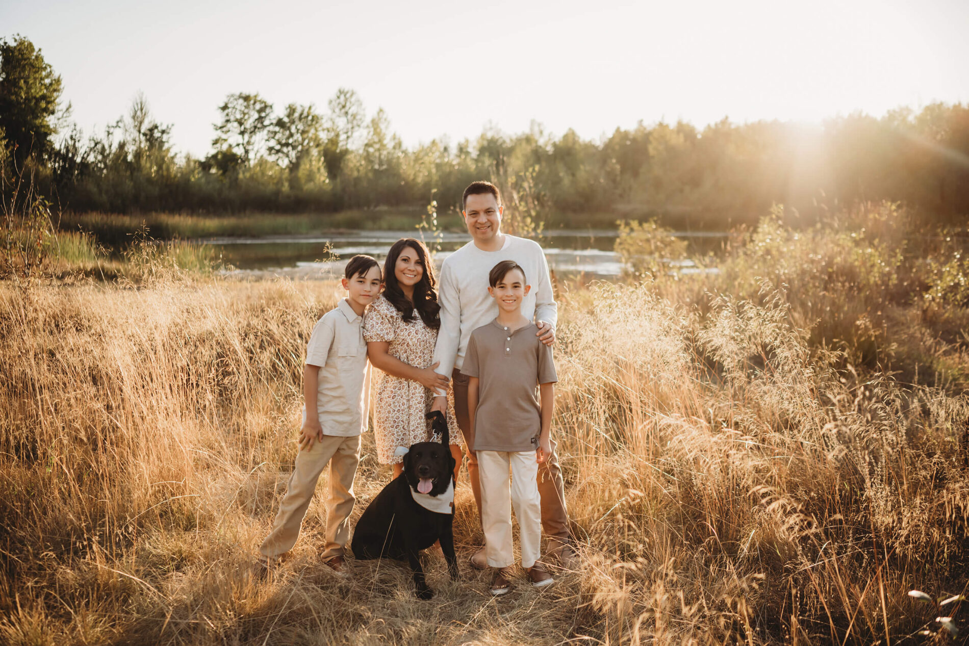 Lifestyle family photo session with mom, dad, two sons, and their dog in a beautiful sunlit field and pond in the background