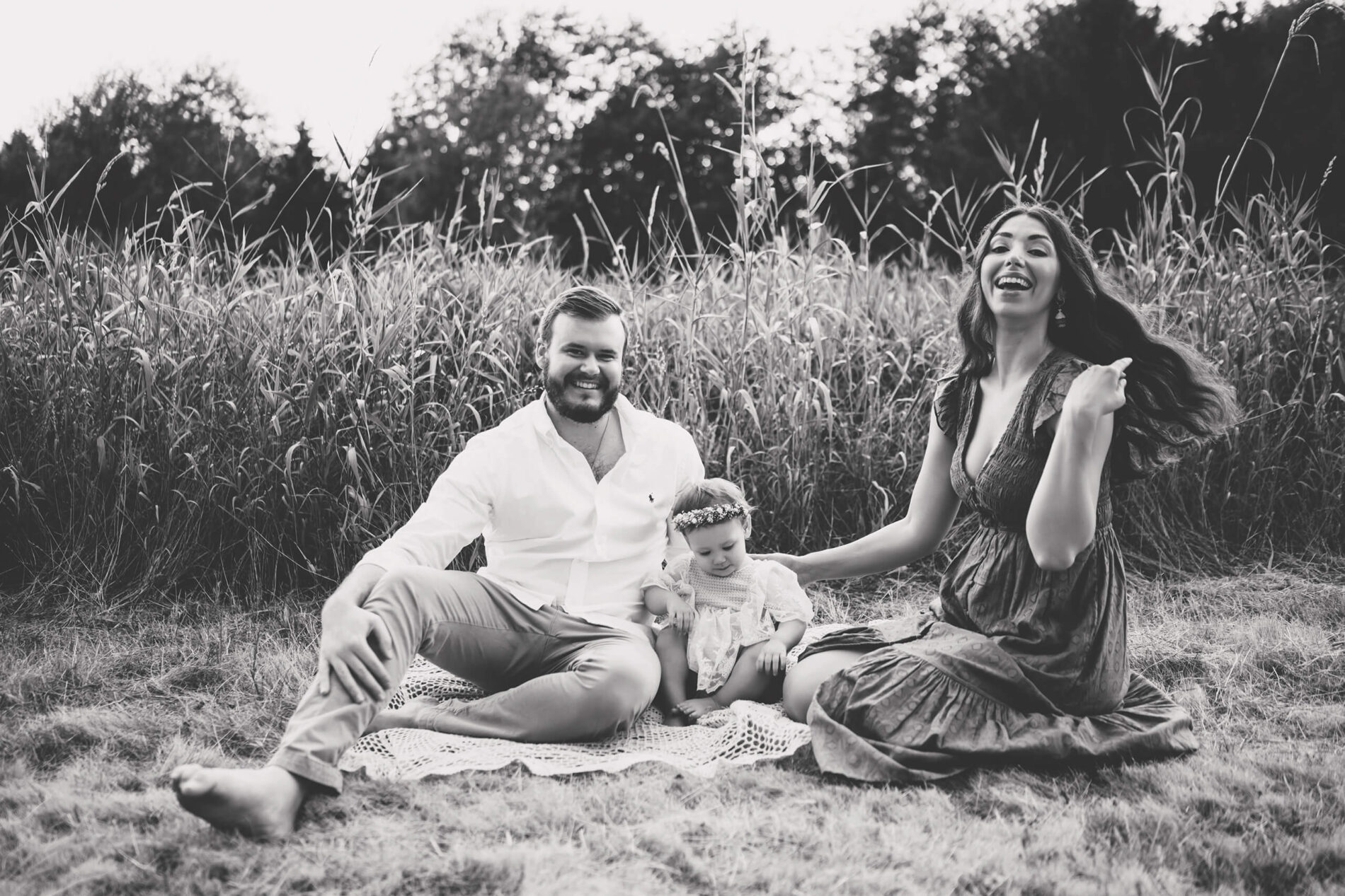 Black and white family lifestyle photo of mom and dad posing with their toddler daughter in a field of tall grass
