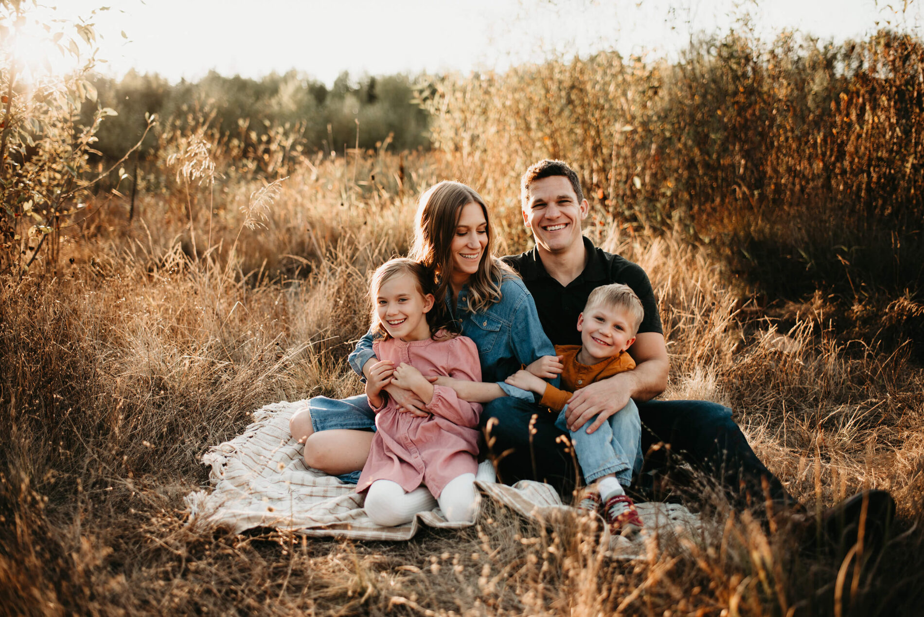Family lifestyle photography with mom, dad, and two children sitting in a filed of tall grass, hugging and smiling