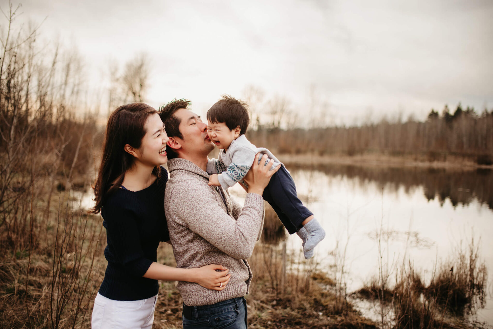 Family lifestyle photo session of two parents playing with their toddler son in a natural setting with a pond in the background