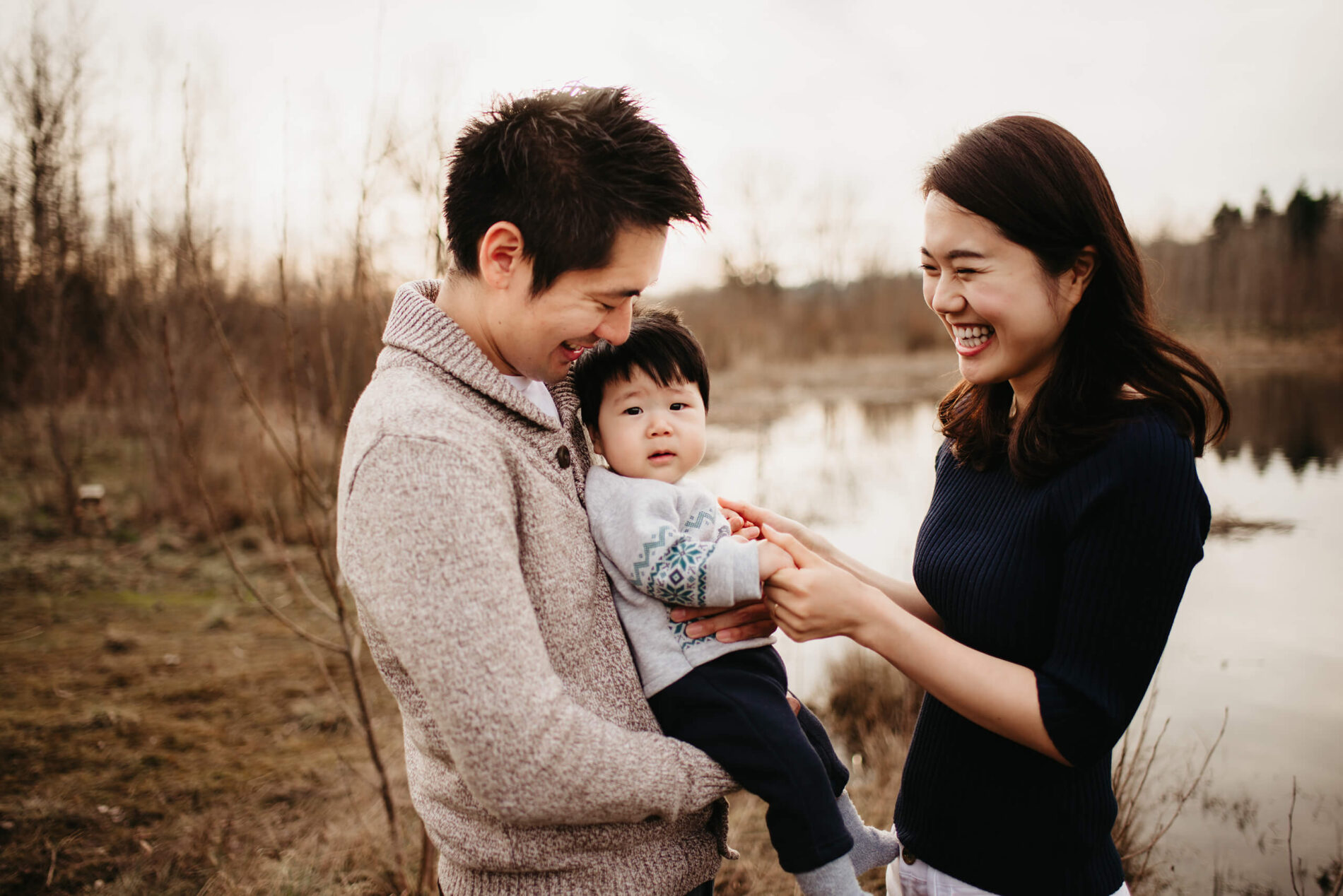 Family photo of a dad holding his toddler son in arms with mom smiling and playing with their son
